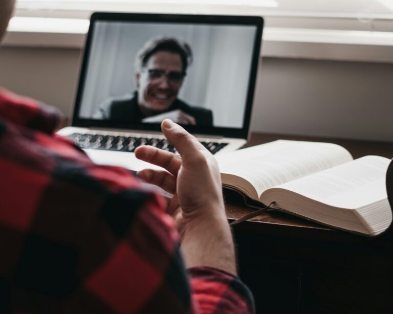 Person on a video call shown on a laptop screen while a book lies open on a desk in the foreground.
