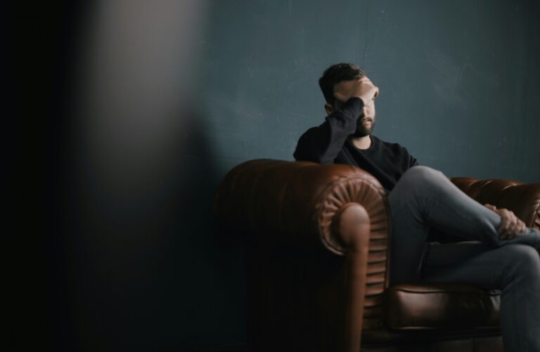 Man in a black sweater sits on a brown leather sofa against a dark teal wall, hand pressed to his forehead in a moment of distress.